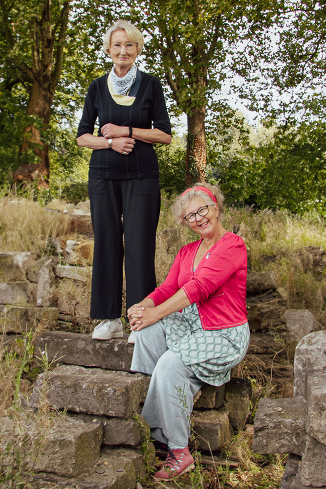 Zwei Frauen vor einer Baumreihe, links stehend, rechts sitzend.