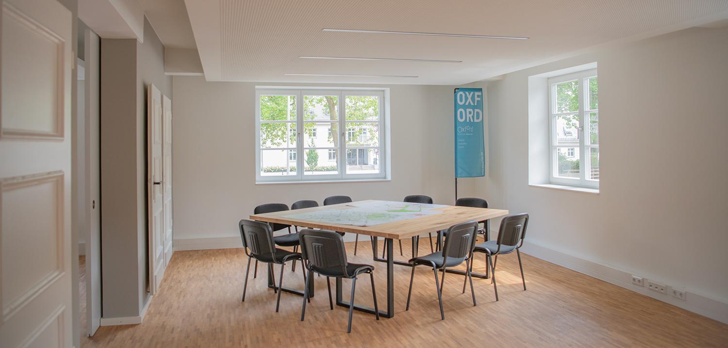 Ein Büroraum mit einer Tischgruppe. In der Ecke eine blaue Fahne mit der Aufschrift Oxford-Quartier.